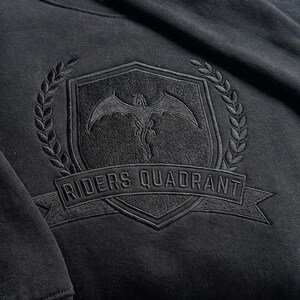 May include: Black sweatshirt with an embroidered emblem. The emblem features a shield with a dragon, surrounded by a laurel wreath, and the text "RIDERS QUADRANT" on a banner. The design is tone-on-tone.