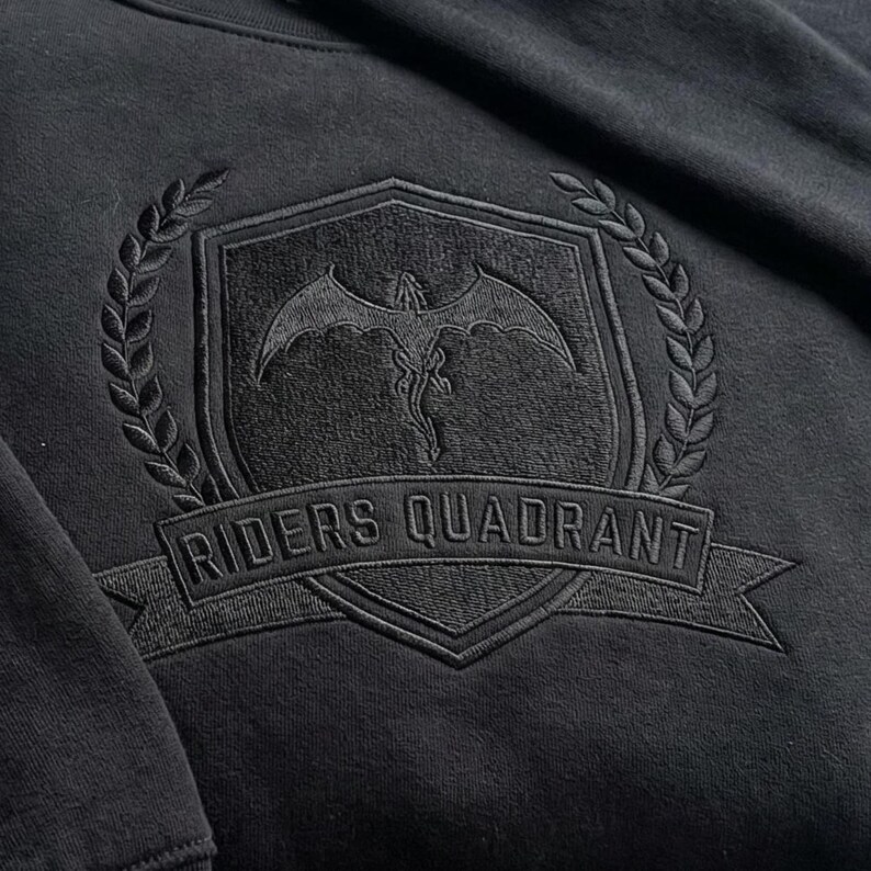 May include: Black sweatshirt with an embroidered emblem. The emblem features a shield with a dragon, surrounded by a laurel wreath, and the text "RIDERS QUADRANT" on a banner. The design is tone-on-tone.