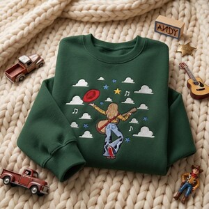 May include: A dark green crewneck sweatshirt featuring an embroidered design of a person with long blonde hair wearing a yellow plaid shirt and blue jeans, holding a guitar and a red hat, surrounded by white clouds, musical notes, and stars.