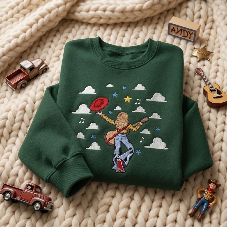 May include: A dark green crewneck sweatshirt featuring an embroidered design of a person with long blonde hair wearing a yellow plaid shirt and blue jeans, holding a guitar and a red hat, surrounded by white clouds, musical notes, and stars.