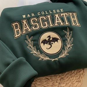 Puede incluir: Una sudadera verde oscuro con el texto "WAR COLLEGE BASGIATH" en una fuente universitaria. Un emblema circular con un dragón negro está centrado debajo del texto, rodeado por una corona de laurel. La sudadera está doblada sobre una superficie tejida.