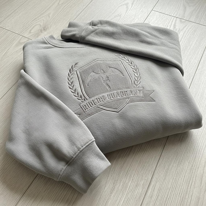 May include: A light gray sweatshirt with the embroidered text "RIDERS QUADRANT" on a shield-shaped emblem. The emblem features a winged figure and is surrounded by a laurel wreath. The sweatshirt is folded on a light wood surface.