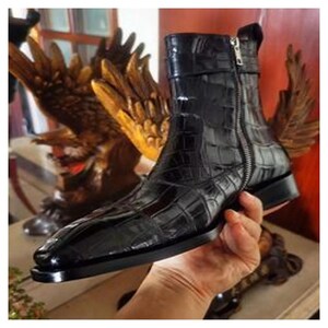 May include: A black leather ankle boot with a crocodile-effect texture. The boot has a side zip, a small strap, and a square toe. The boot is held by a hand, highlighting its shiny finish and design. The boot's design is very stylish.