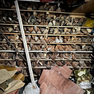 May include: A workshop storage area filled with wooden shoe lasts on shelves. Various sizes and shapes of lasts are visible, along with leather hides and other shoemaking materials. A wooden support beam is in the foreground.