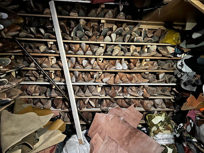 May include: A workshop storage area filled with wooden shoe lasts on shelves. Various sizes and shapes of lasts are visible, along with leather hides and other shoemaking materials. A wooden support beam is in the foreground.