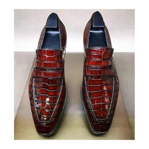 Op de afbeelding: Een paar roodbruine leren loafers met een krokodillenleertextuur. De schoenen hebben een vierkante neus en een decoratieve band over de bovenkant. De zolen zijn gestikt met een zilveren draad.