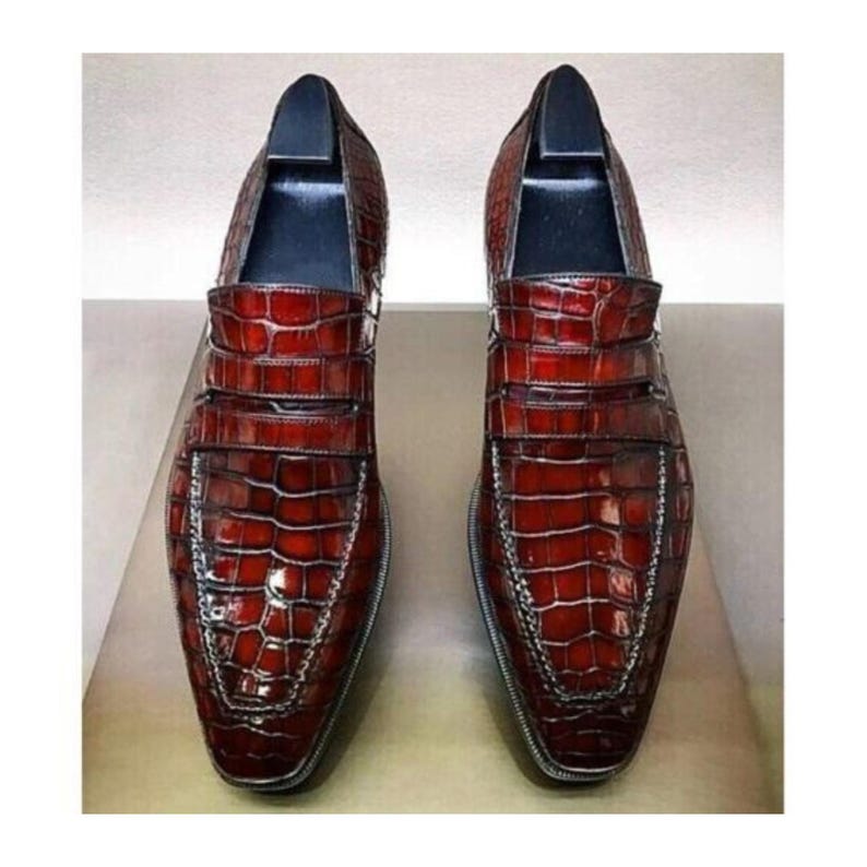 Op de afbeelding: Een paar roodbruine leren loafers met een krokodillenleertextuur. De schoenen hebben een vierkante neus en een decoratieve band over de bovenkant. De zolen zijn gestikt met een zilveren draad.
