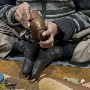 May include: A person seated cross-legged, working on a brown leather shoe. The artisan holds the shoe's upper, showcasing the craftsmanship. The scene includes tools and materials, highlighting the shoemaking process.