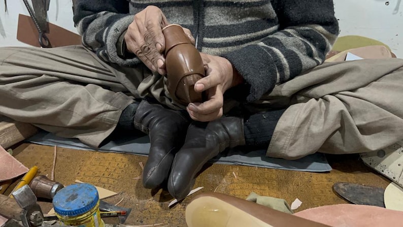 May include: A person seated cross-legged, working on a brown leather shoe. The artisan holds the shoe's upper, showcasing the craftsmanship. The scene includes tools and materials, highlighting the shoemaking process.