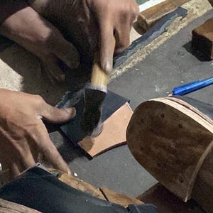 May include: A close-up shot of shoemaking. Hands are shown working on leather and shoe components. Tools like brushes, knives, and a pen are visible. The focus is on the craftsmanship of shoe construction.