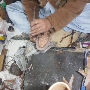 May include: A shoemaker working on a light brown leather shoe. The shoemaker is using a tool to shape the shoe. Various tools and leather scraps are scattered around the workspace. The shoe is in the early stages of construction.