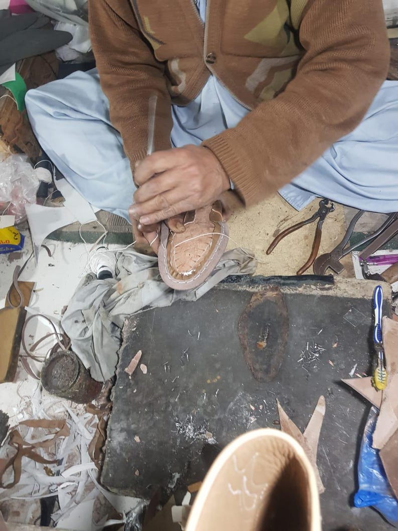 May include: A shoemaker working on a light brown leather shoe. The shoemaker is using a tool to shape the shoe. Various tools and leather scraps are scattered around the workspace. The shoe is in the early stages of construction.