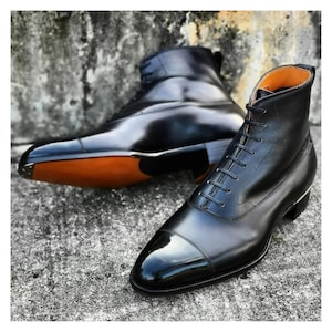 May include: Two black leather dress boots with a high-shine finish. The boots have a classic lace-up design and a cap toe. The soles are a contrasting brown color. The boots are set against a textured gray background.