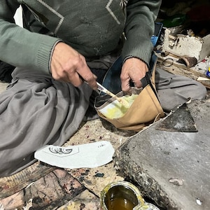 May include: A person is working on a shoe, applying adhesive with a small tool. The shoe is in the process of being made. The person is wearing a green sweater and gray pants. A shoe sole is visible.