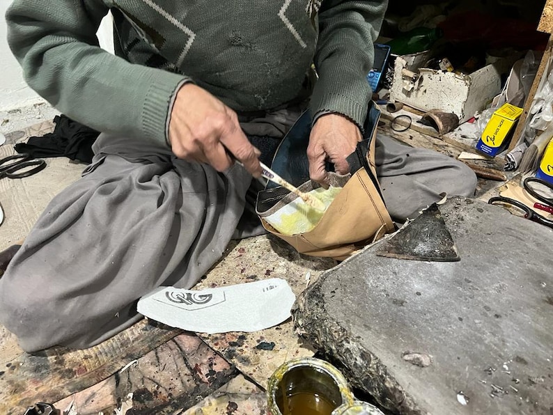 May include: A person is working on a shoe, applying adhesive with a small tool. The shoe is in the process of being made. The person is wearing a green sweater and gray pants. A shoe sole is visible.