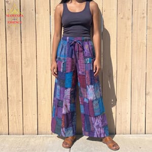 May include: Patchwork wide-leg trousers in purple, blue, and teal hues. The trousers have a drawstring waist and a mix of fabric patterns. The model is wearing a dark grey vest top and brown sandals.