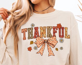 Könnte beinhalten: Ein cremefarbenes Langarm-Sweatshirt mit dem Wort THANKFUL in einer Mischung aus Karomustern und Leopardenmuster. Unter dem Text befindet sich eine große Schleife mit Leopardenmuster, umgeben von kleinen weißen Gänseblümchen und Herbstblättern.