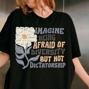 Op de afbeelding: Zwart T-shirt met een bloemenafbeelding en de tekst "IMAGINE BEING AFRAID OF DIVERSITY BUT NOT DICTATORSHIP". Het ontwerp toont een witte bloem met prikkeldraad en grijze bladeren.