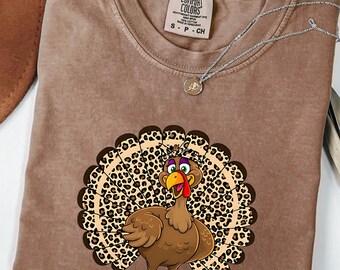 Könnte beinhalten: Ein braunes T-Shirt mit kurzen Ärmeln und einer Grafik eines Truthahns mit einem Schwanz im Leopardenmuster. Das T-Shirt hat einen Rundhalsausschnitt und ein aufgedrucktes Comfort Colors-Markenetikett am Innenkragen. Der Truthahn hat braune Federn.