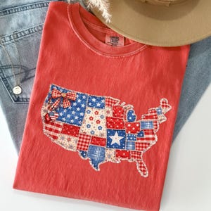 May include: A red Comfort Colors t-shirt with a patchwork design of the United States map in patriotic colours. The map includes bow and star accents. The shirt is paired with blue jeans and a tan hat.