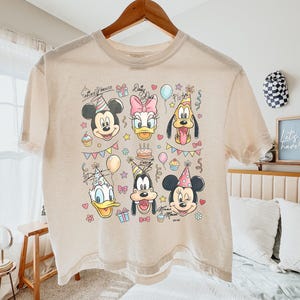Op de afbeelding: Een beige t-shirt met een kleurrijke afbeelding van Mickey Mouse, Daisy Duck, Pluto, Donald Duck, Goofy en Minnie Mouse, elk met een feesthoed. Het ontwerp bevat cupcakes, ballonnen en een taart, met de woorden "Mickey Mouse" en "Daisy" in script.
