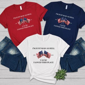 May include: Three t-shirts in red, white, and navy blue, each with the text "I'M JUST MAD AS HELL CAUSE I LOVED THIS PLACE" and two American flags. The shirts are paired with blue denim jeans. A small green plant is visible.