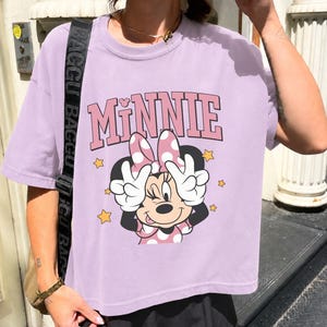 Pode incluir: Camiseta lil&aacute;s claro com um gr&aacute;fico da Minnie Mouse. O design inclui a palavra "MINNIE" em rosa, Minnie Mouse com um la&ccedil;o rosa e estrelas amarelas. Minnie faz um sinal de paz com as m&atilde;os.