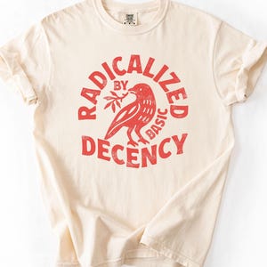 K&ouml;nnte beinhalten: Cremefarbenes T-Shirt mit dem roten Text "RADICALIZED BY BASIC DECENCY" in einem kreisf&ouml;rmigen Design. Ein roter Vogel, der einen Zweig h&auml;lt, befindet sich in der Mitte. Das Shirt hat hochgekrempelte &Auml;rmel und ein Etikett am Halsausschnitt.