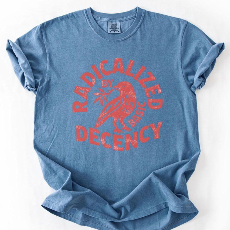 K&ouml;nnte beinhalten: Blaues T-Shirt mit Rundhalsausschnitt und kurzen &Auml;rmeln. Das Shirt hat eine rote Grafik in einem kreisf&ouml;rmigen Design mit der Aufschrift "RADICALIZED BY BASIC DECENCY" und einer Vogelillustration in der Mitte.