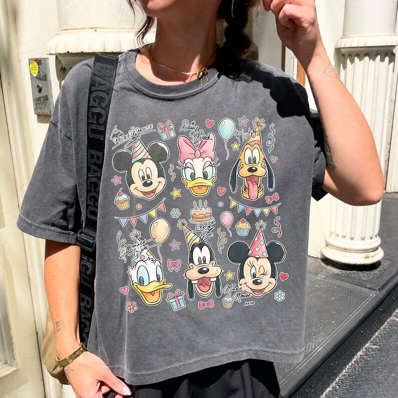 Op de afbeelding: Antracietgrijs T-shirt met een kleurrijke afbeelding van Disney-figuren, waaronder Mickey Mouse, Minnie Mouse, Donald Duck, Daisy Duck, Goofy en Pluto, die feesthoeden dragen en omringd zijn door verjaardagsversieringen.