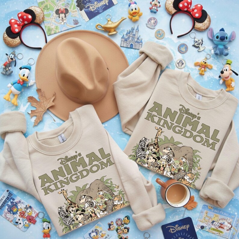 May include: Two beige sweatshirts with the text "Disney's Animal Kingdom" and a graphic of Disney characters and animals. The image also includes Disney-themed accessories like headbands, keychains, and a hat, all arranged on a light blue surface.