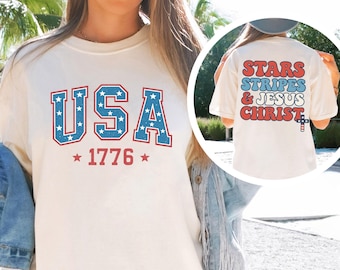 Puede incluir: Camiseta color crema con la palabra "USA" en letras azules con estrellas blancas y el año "1776" en rojo. La parte trasera de la camiseta presenta las palabras "Stars, Stripes & Jesus Christ" en rojo y azul.