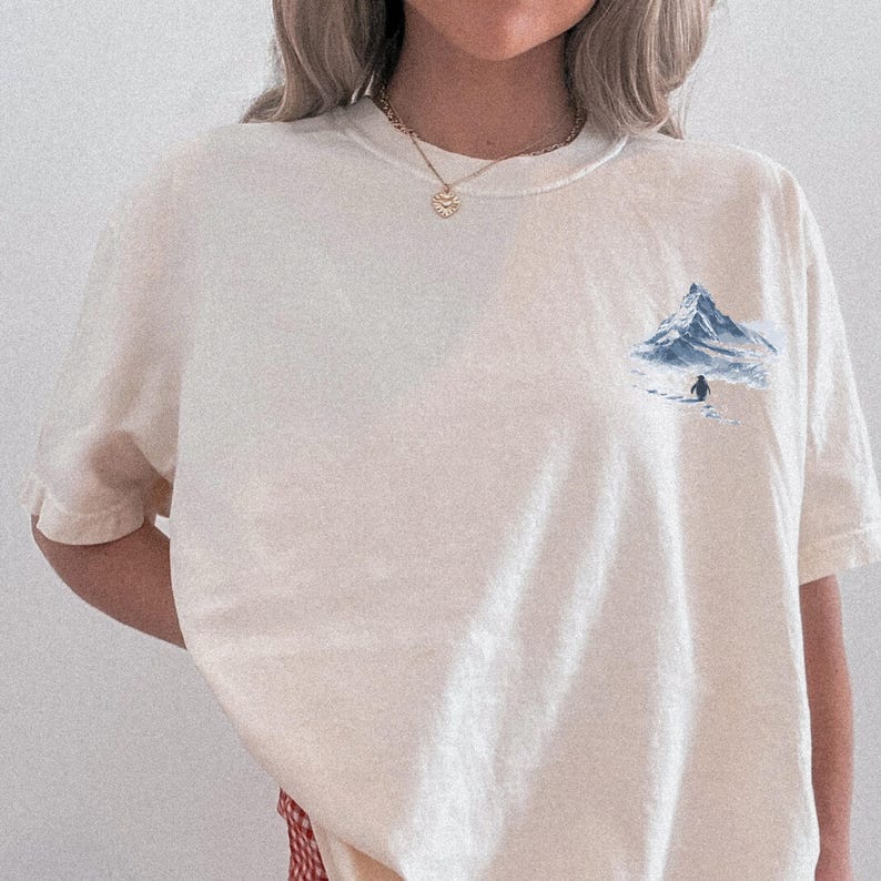 Puede incluir: Camiseta color crema con un peque&ntilde;o gr&aacute;fico de una monta&ntilde;a azul y un ping&uuml;ino. La camiseta tiene mangas cortas y cuello redondo. El dise&ntilde;o est&aacute; en el &aacute;rea del pecho izquierdo.