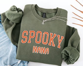 May include: An olive green crewneck sweatshirt featuring the text SPOOKY NANA in orange varsity-style lettering with a distressed texture. The sweatshirt has long sleeves with ribbed cuffs and a black neck label, paired with blue denim jeans.
