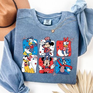 May include: A blue crew neck sweatshirt featuring six Disney characters in patriotic outfits. Donald Duck, Mickey Mouse, Daisy Duck, Pluto, and Minnie Mouse are depicted in red, white, and blue designs. A gold necklace and a brown hat are also visible.