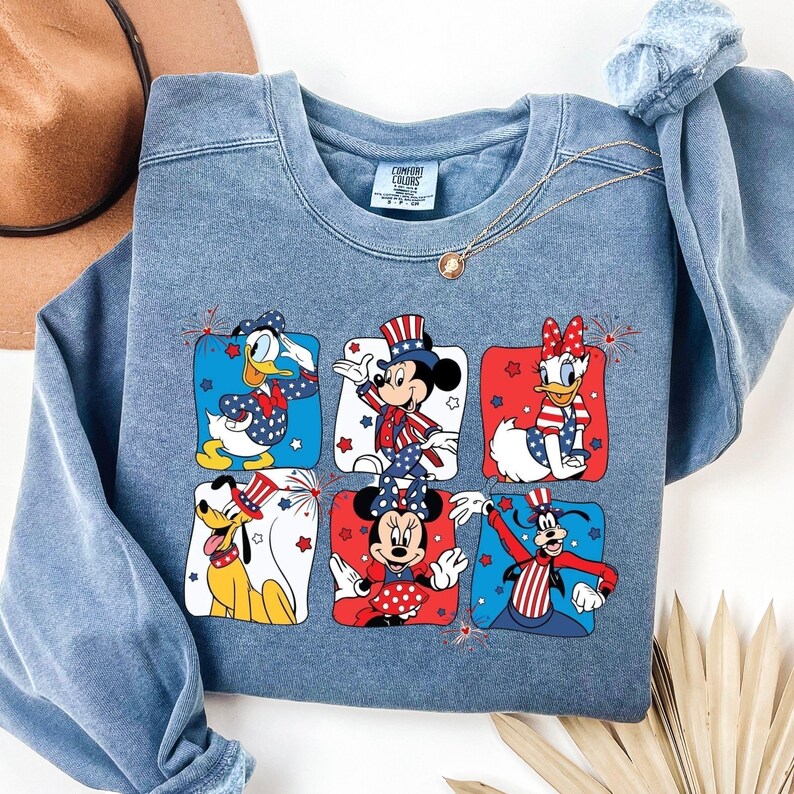 May include: A blue crew neck sweatshirt featuring six Disney characters in patriotic outfits. Donald Duck, Mickey Mouse, Daisy Duck, Pluto, and Minnie Mouse are depicted in red, white, and blue designs. A gold necklace and a brown hat are also visible.