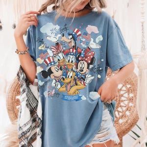 May include: A light blue t-shirt featuring a graphic of Disney characters Mickey Mouse, Minnie Mouse, Donald Duck, Daisy Duck, Goofy, and Pluto in patriotic attire. The shirt has the text "America 250th USA Anniversary 1776-2026".