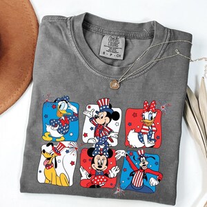 May include: Gray Comfort Colors t-shirt featuring six Disney characters in patriotic outfits. Donald Duck, Mickey Mouse, Daisy Duck, Minnie Mouse, Goofy, and Pluto are depicted in red, white, and blue designs. A silver necklace with a pendant is draped across the shirt.