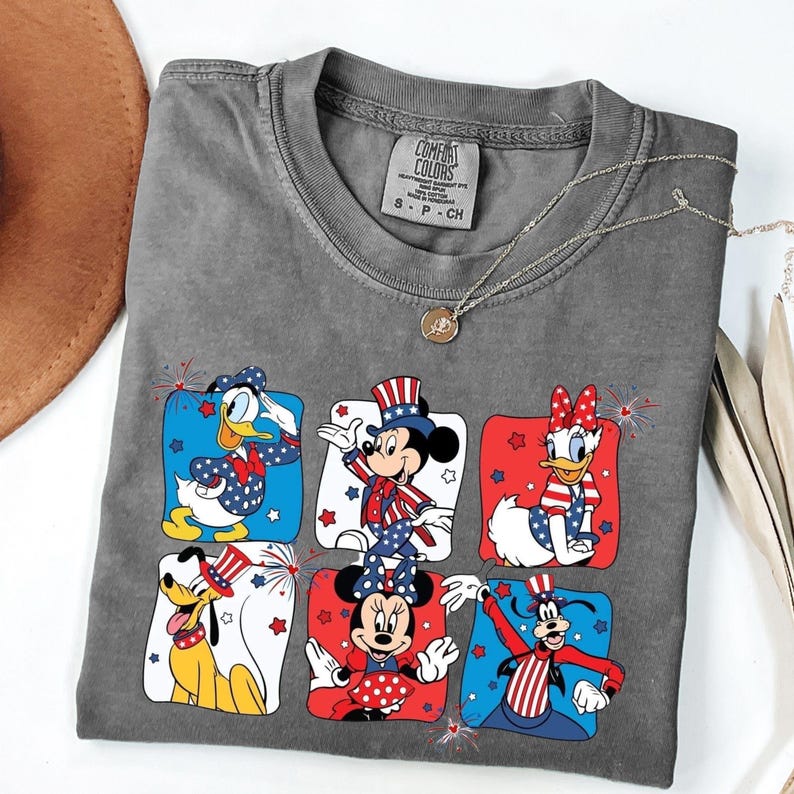 May include: Gray Comfort Colors t-shirt featuring six Disney characters in patriotic outfits. Donald Duck, Mickey Mouse, Daisy Duck, Minnie Mouse, Goofy, and Pluto are depicted in red, white, and blue designs. A silver necklace with a pendant is draped across the shirt.