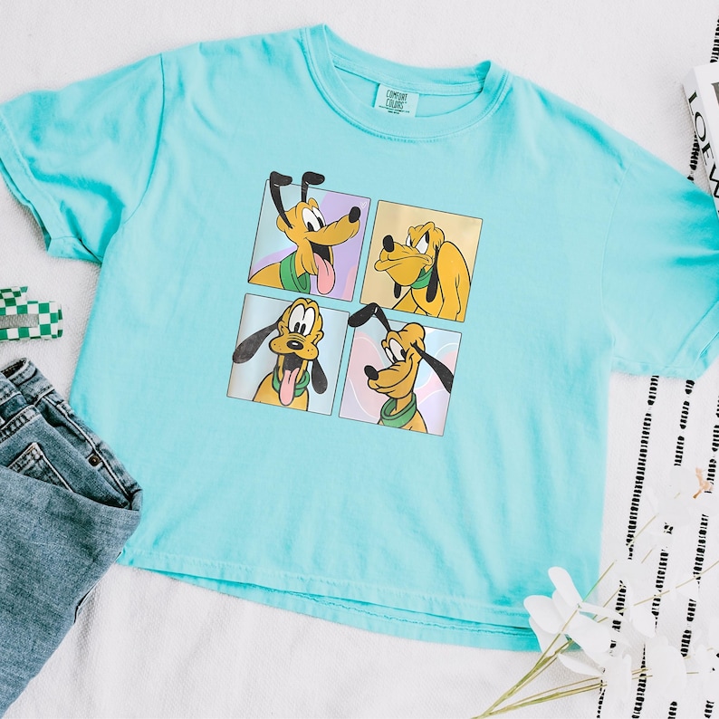 May include: A light blue t-shirt featuring a four-panel graphic of Pluto, the Disney cartoon dog. Each panel shows Pluto with a different expression. The shirt is paired with blue jeans and a small green and white checkered item.