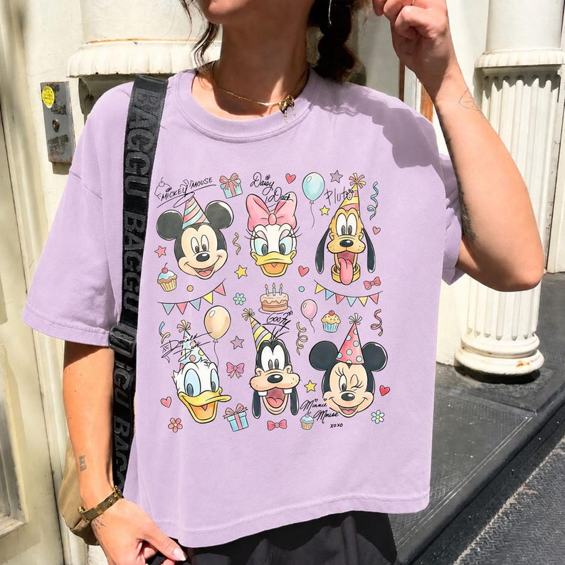 Op de afbeelding: Een lichtpaars T-shirt met een kleurrijke afbeelding van Mickey Mouse, Daisy Duck, Pluto, Donald Duck, Goofy en Minnie Mouse met feesthoeden. Het ontwerp bevat cupcakes, ballonnen en confetti. Het T-shirt heeft een relaxte pasvorm.