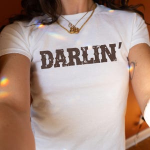 May include: White t-shirt with the word "DARLIN'" printed in brown, distressed lettering. The shirt has a crew neck and short sleeves. A gold necklace is worn around the neck.