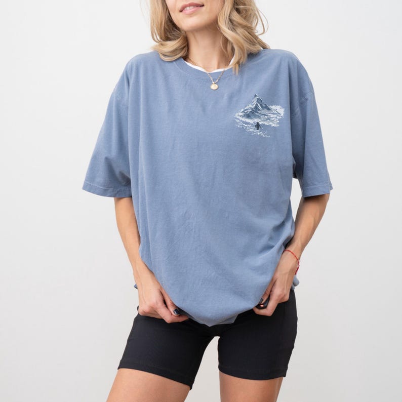 Puede incluir: Camiseta azul claro con cuello redondo blanco y un gr&aacute;fico de monta&ntilde;a en la parte superior izquierda del pecho. El gr&aacute;fico representa una cordillera con una persona. La camiseta se combina con pantalones cortos negros.