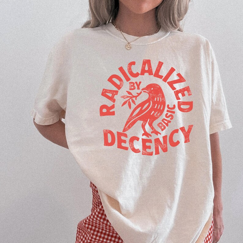 K&ouml;nnte beinhalten: Cremefarbenes T-Shirt mit einem roten, verwaschenen Aufdruck. Der Aufdruck zeigt einen Vogel mit einem Zweig, umgeben von den Worten "RADICALIZED BY BASIC DECENCY" in einem kreisf&ouml;rmigen Layout. Das Shirt hat kurze &Auml;rmel und eine lockere Passform.