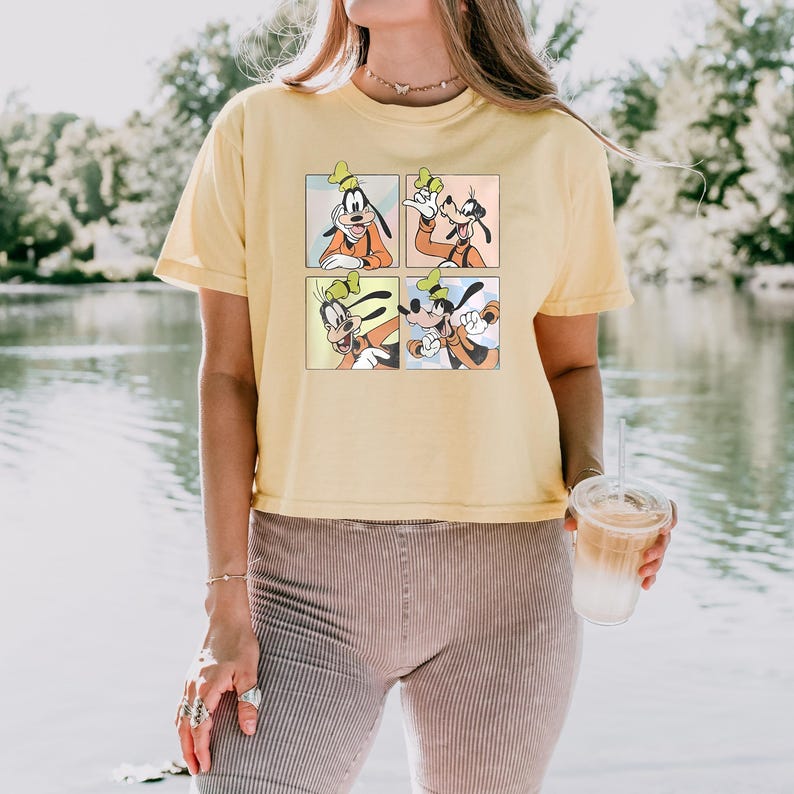 May include: A light yellow t-shirt featuring a four-panel graphic of Goofy in various poses. Goofy is wearing an orange shirt, green hat, and black ears. The shirt is a casual style.