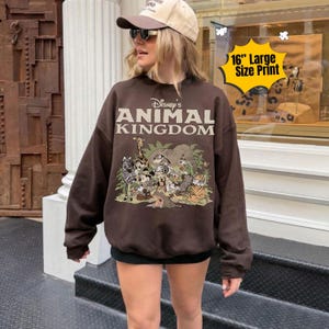 May include: A brown sweatshirt featuring the text "Disney's Animal Kingdom" above a graphic of various cartoon animals. The sweatshirt has a large 16-inch print and is worn by a person wearing a baseball cap and sunglasses.