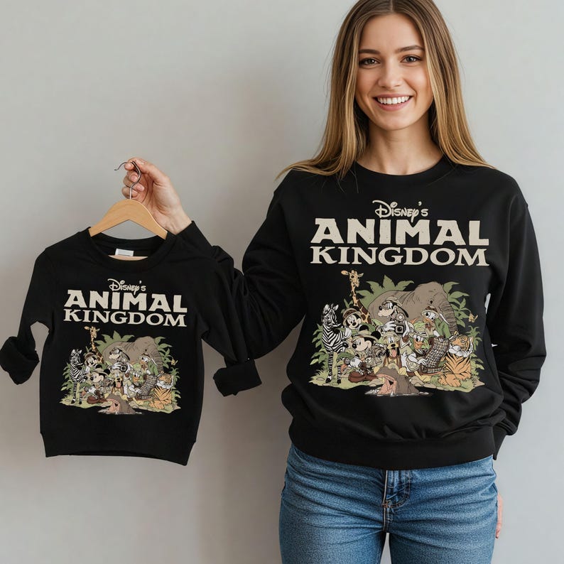 May include: Black sweatshirts featuring the text "Disney's Animal Kingdom" in white. The sweatshirts display a colorful graphic of various animals. One sweatshirt is held up by a person, and the other is being worn.
