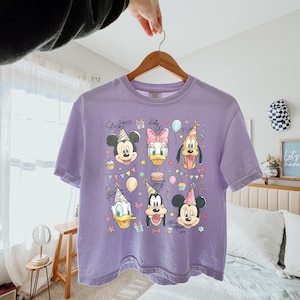 Op de afbeelding: Een lavendelkleurig T-shirt met een kleurrijke afbeelding van Mickey Mouse, Minnie Mouse, Donald Duck, Daisy Duck, Goofy en Pluto, allemaal met feesthoeden. Het ontwerp bevat ballonnen, cupcakes en de tekst "Mickey & Friends".