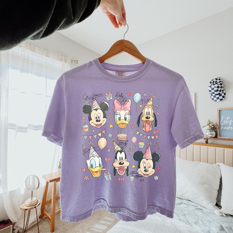 Op de afbeelding: Een lavendelkleurig T-shirt met een kleurrijke afbeelding van Mickey Mouse, Minnie Mouse, Donald Duck, Daisy Duck, Goofy en Pluto, allemaal met feesthoeden. Het ontwerp bevat ballonnen, cupcakes en de tekst "Mickey & Friends".