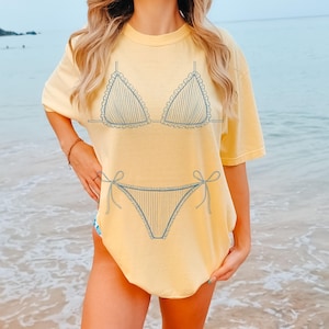 May include: Pale yellow t-shirt with a blue outline of a bikini. The bikini design includes vertical stripes and frilly edges. The t-shirt has a loose fit, suitable for beach outings or everyday wear.